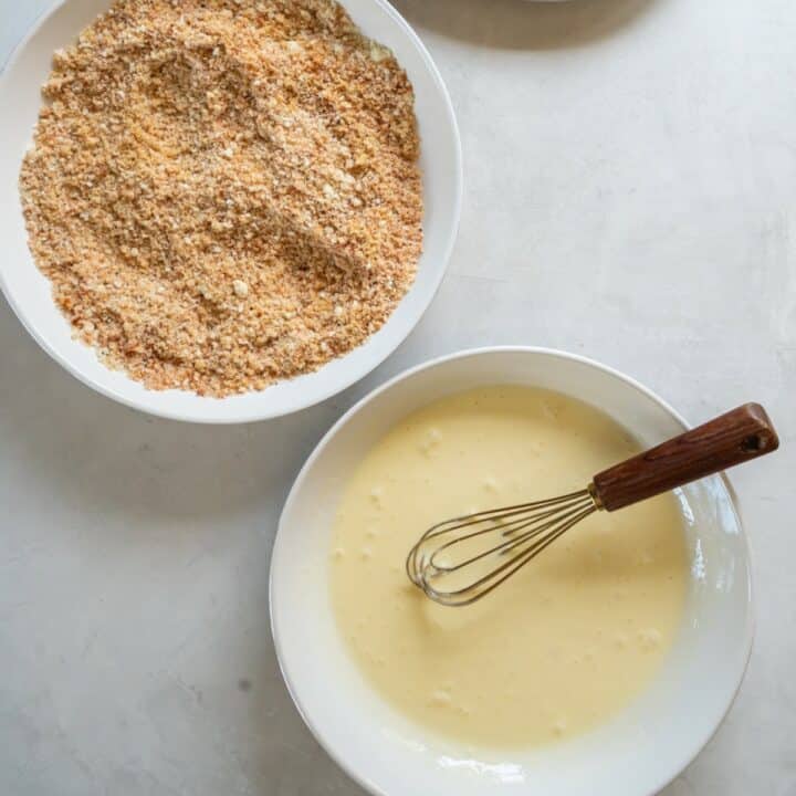 Two bowls for dredging chicken. One with whisked eggs and cream, the other with pork rinds, almond flour, cheese and seasonings.