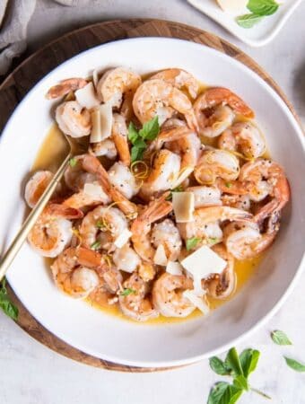 A bowl filled with shrimp scampi in a white wine garlic butter sauce garnished with fresh basil leaves and sliced pieces of fresh Parmesan cheese.