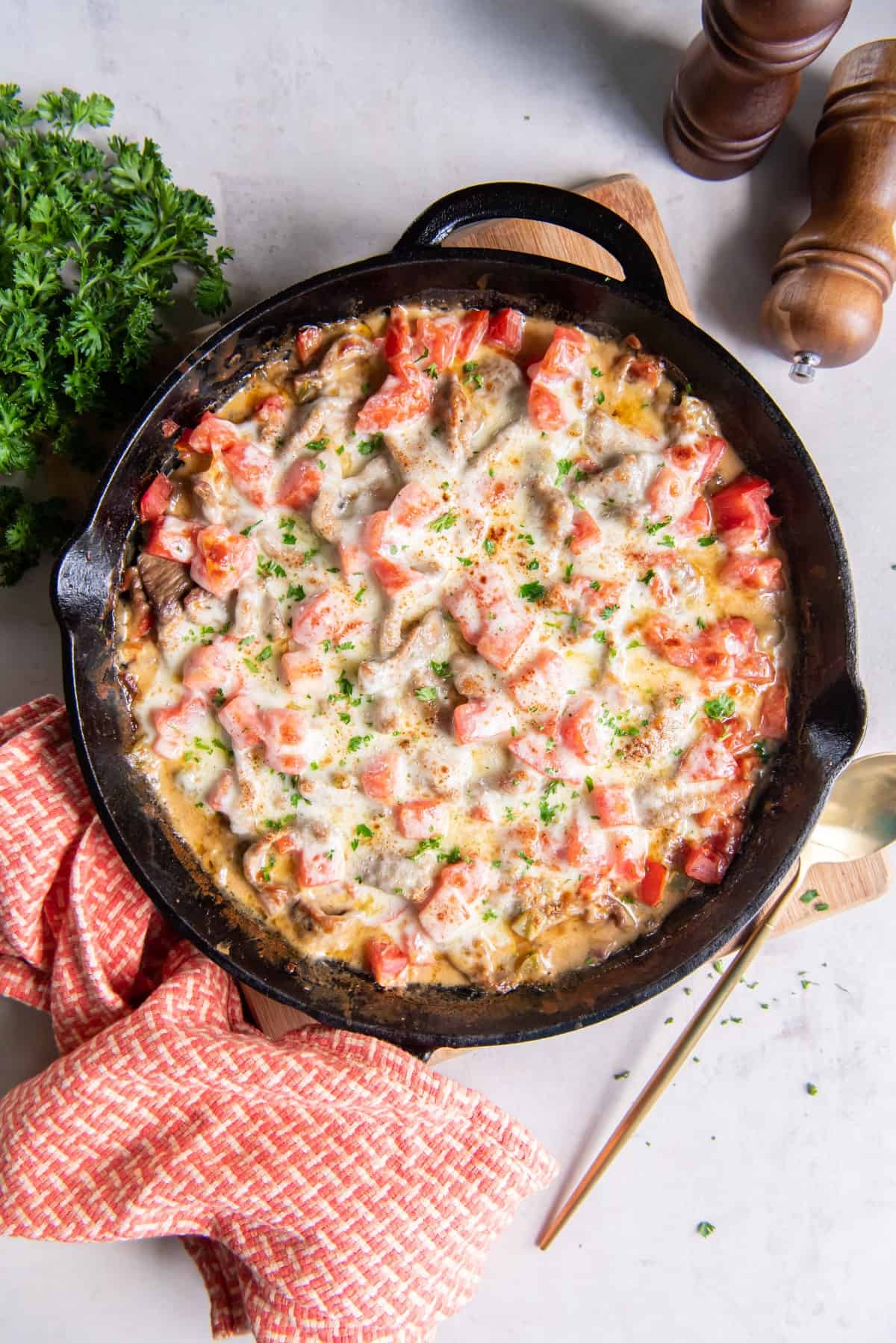 A cast iron pan with baked Philly cheesesteak with steak, cheese and tomatoes, placed on a light wood cutting board with a brass serving utensil and a red and white textured kitchen towel.