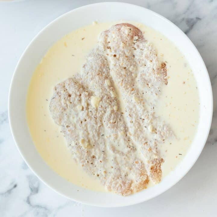 A breadcrumb coated chicken breast being dunked into a bowl of heavy cream.