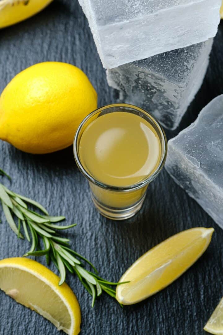 Green tea shot in a shot glass surrounded by slices of lemon and large blocks of ice.