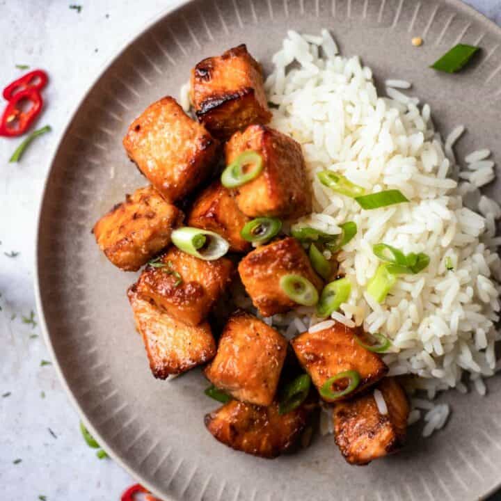 Cooked honey garlic salmon bites on a plate with rice topped with chopped scallions.