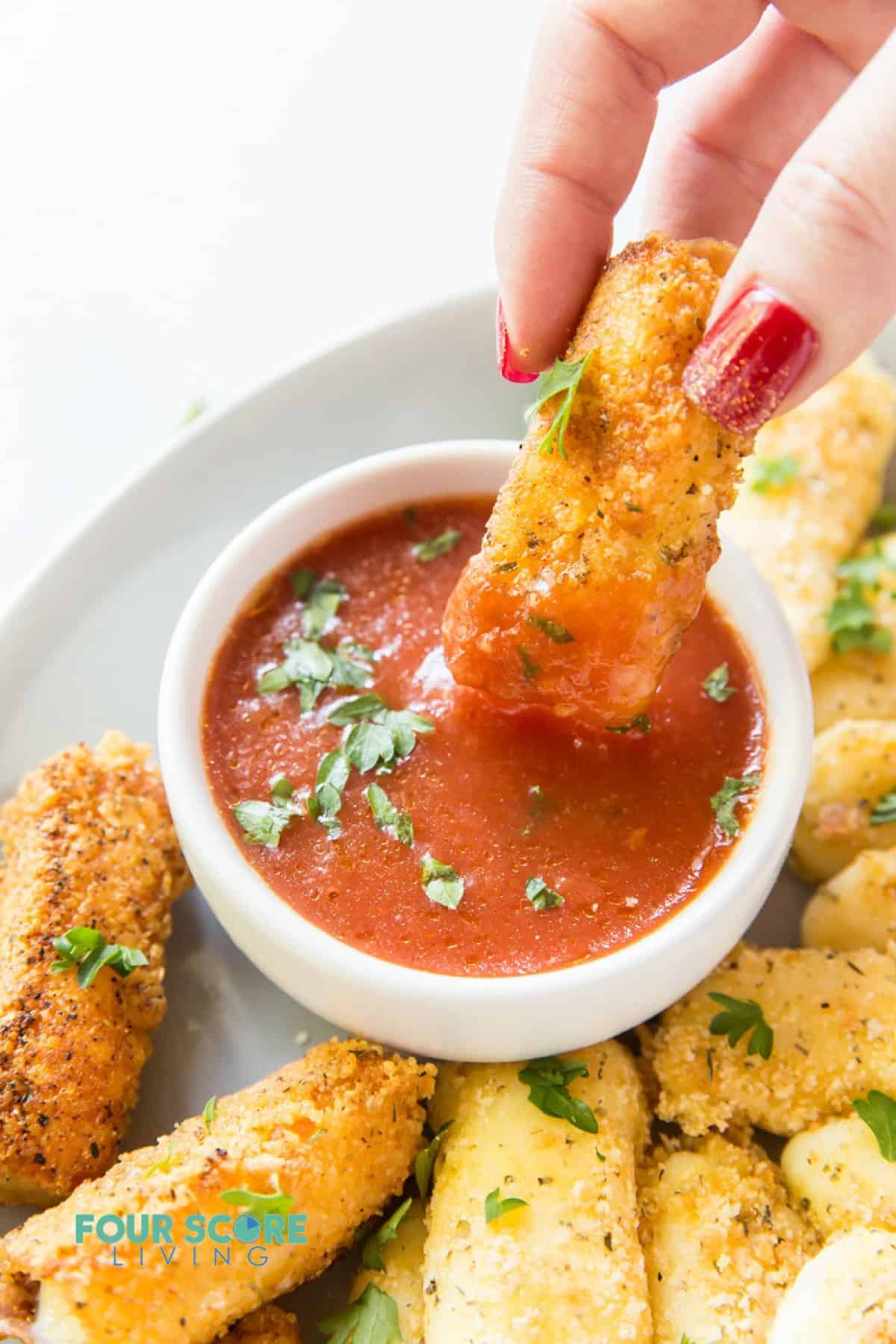 Mozzarella stick being dipped into a small bowl of marinara sauce by a feminine hand.