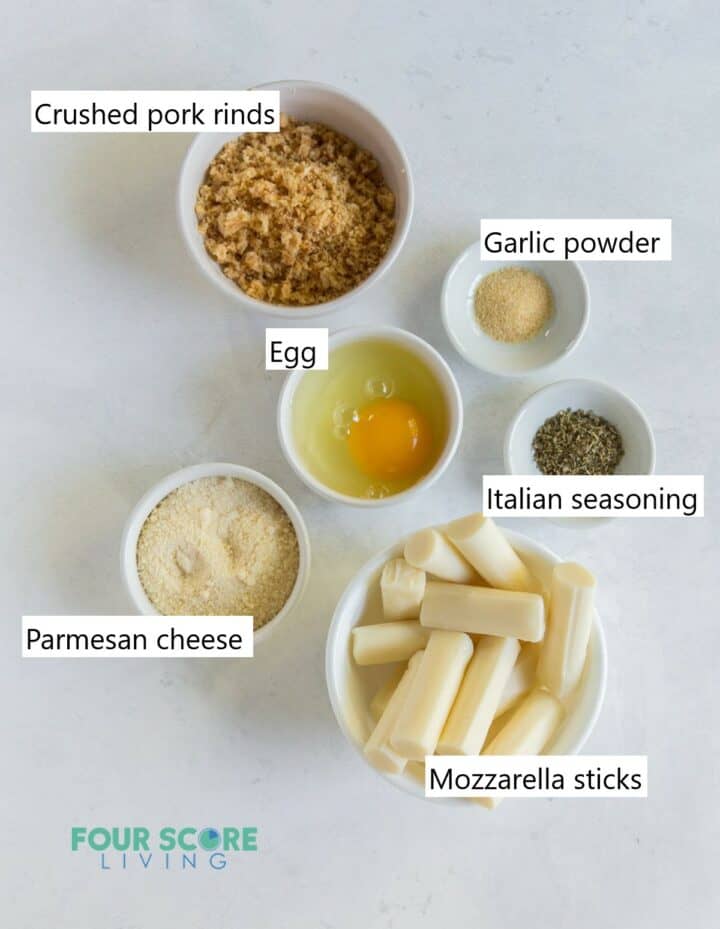 Ingredients for making keto mozzarella sticks, each in separate bowls. Including cheese sticks, pork rinds, egg, and seasoning.