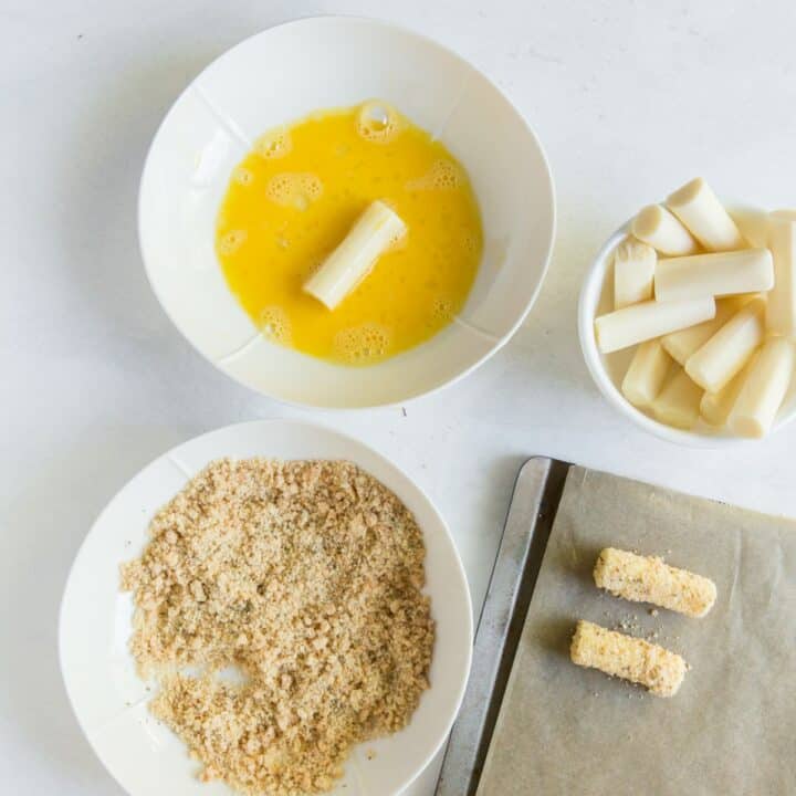 A mozzarella stick in the egg wash bowl. There are also two fully breaded sticks on a parchment lined baking sheet.