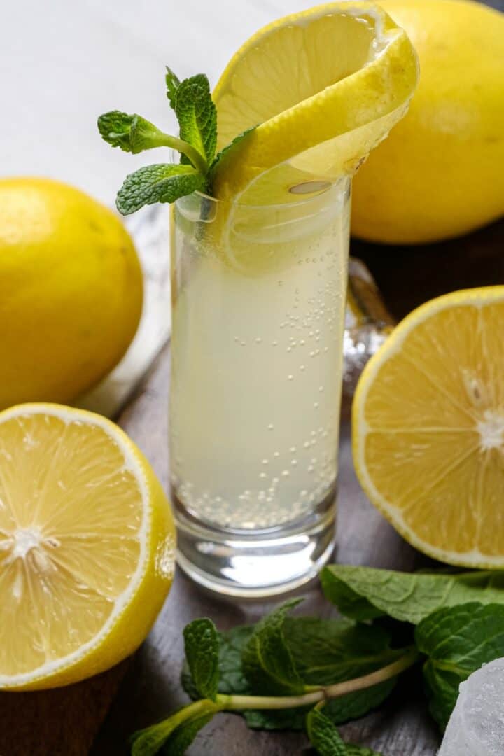 White Tea Shot in a wooden cutting board. There is a slice of lemon and sprig of mint in the shot, and the glass is surrounded by mint, lemons, and ice.