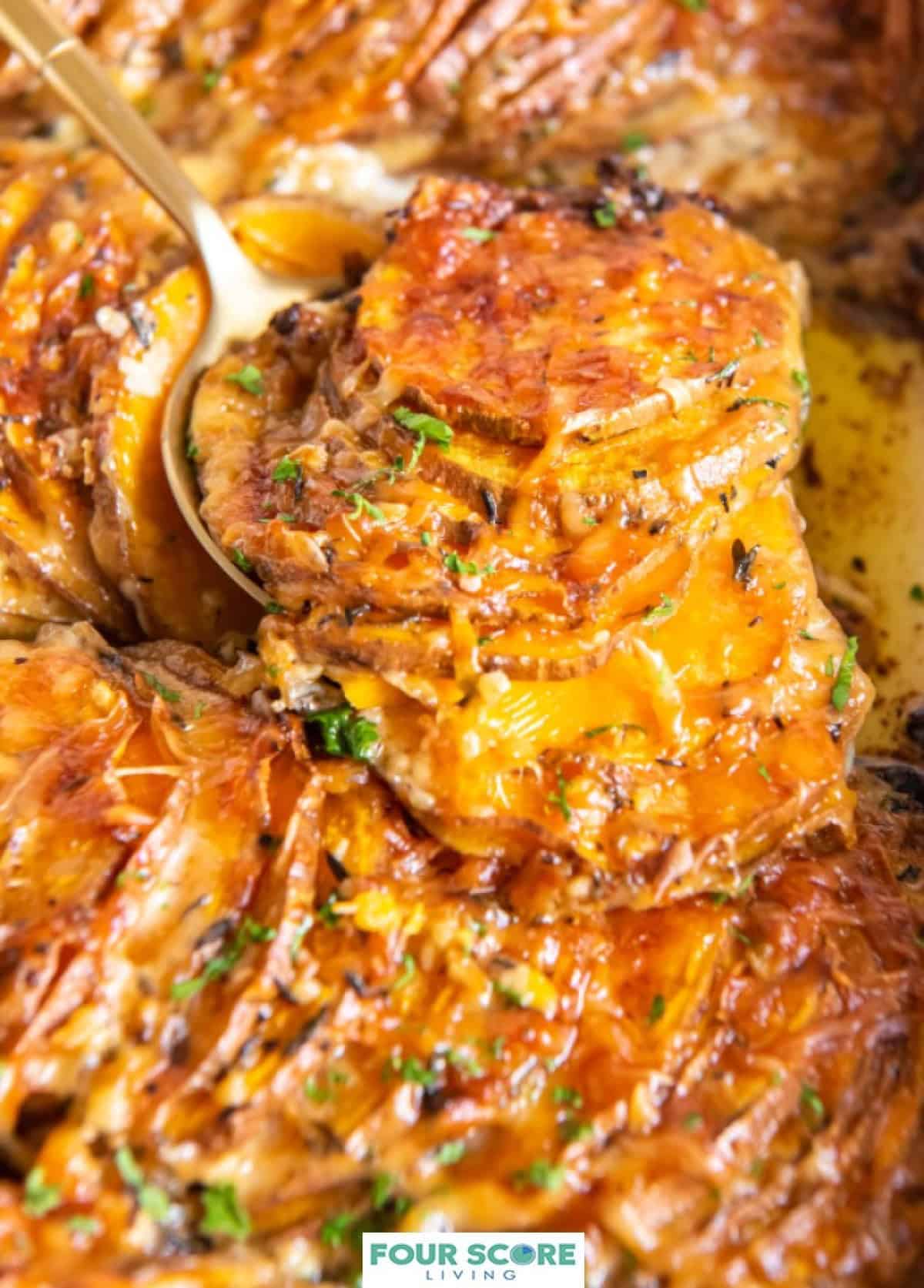 Scalloped sweet potatoes covered in melted cheese with a sprinkle of fresh green herbs being scooped with a serving utensil.