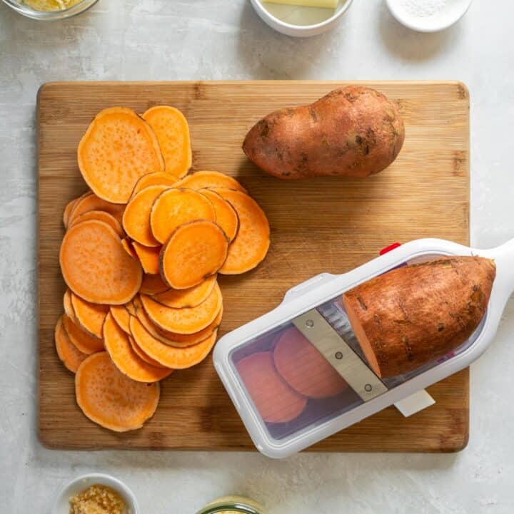 A sweet potato being sliced with a mandoline on a wooden cutting board.