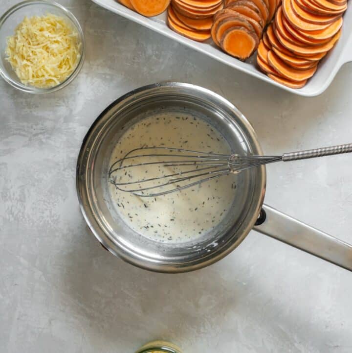 A saucepan with heavy cream being whisked into melted butter, garlic, thyme, salt, pepper and nutmeg.