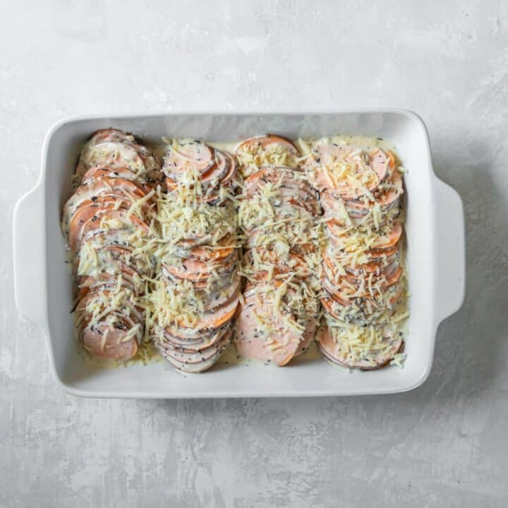 A fully assembled scalloped sweet potato with gruyere cheese sprinkled on top ready to be baked.