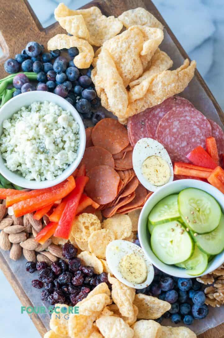 A fully assembled keto charcuterie board with pork rinds, blueberries, salami, pepperoni, ham, almonds, walnuts, pickles, peppers, cheese crisps, crumbled blue cheese and hard boiled eggs.