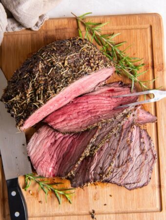 An herb crusted, juicy and tender beef bottom round roast partially sliced on a wood cutting board with a spring of fresh rosemary and a two pronged serving tool and a sharp knife.