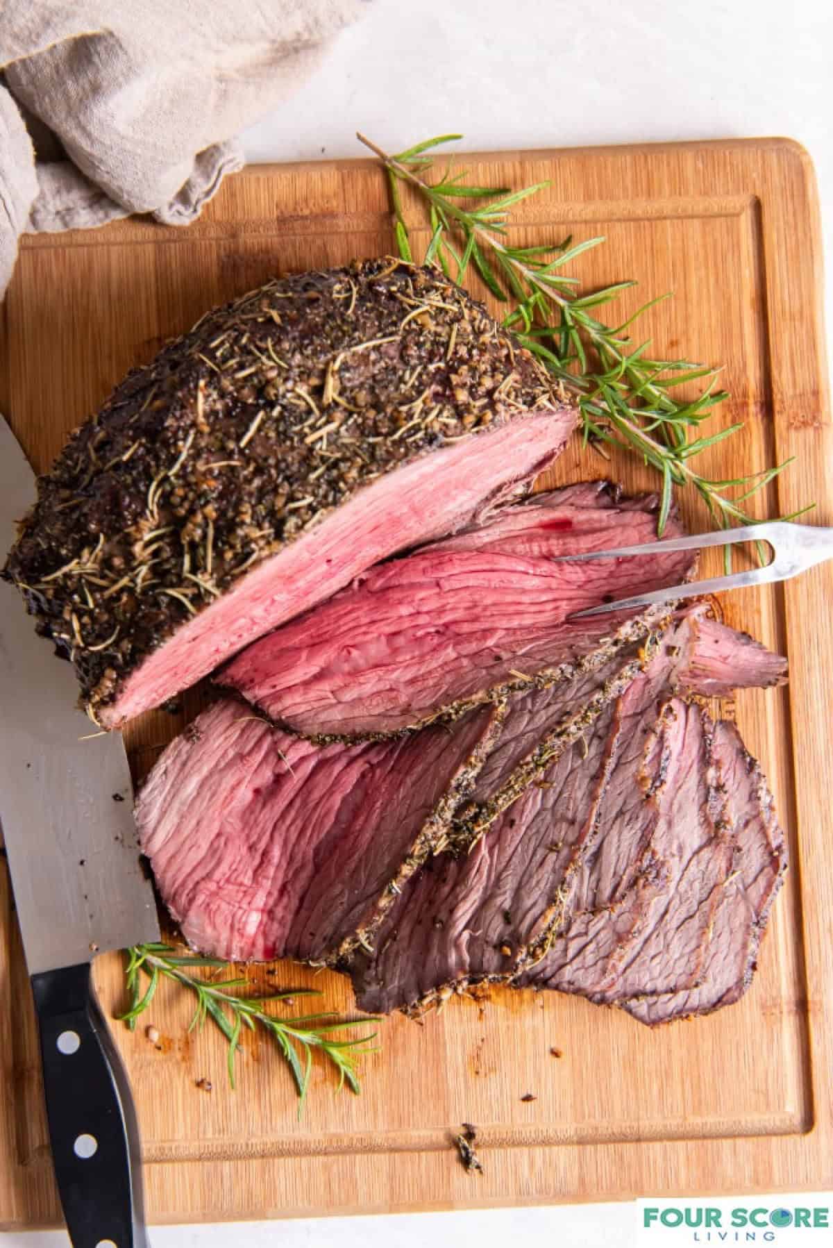 An herb crusted, juicy and tender beef bottom round roast partially sliced on a wood cutting board with a spring of fresh rosemary and a two pronged serving tool and a sharp knife.