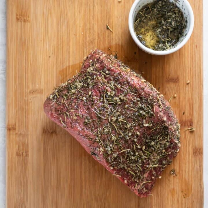 A bottom round roast heavily coated in a herb seasoning mixture on a wooden cutting board.