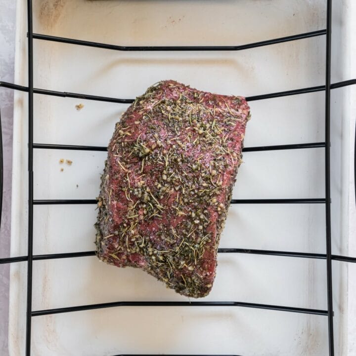 An herb coated beef bottom round roast on a rack in a roasting pan ready to be placed into an oven.