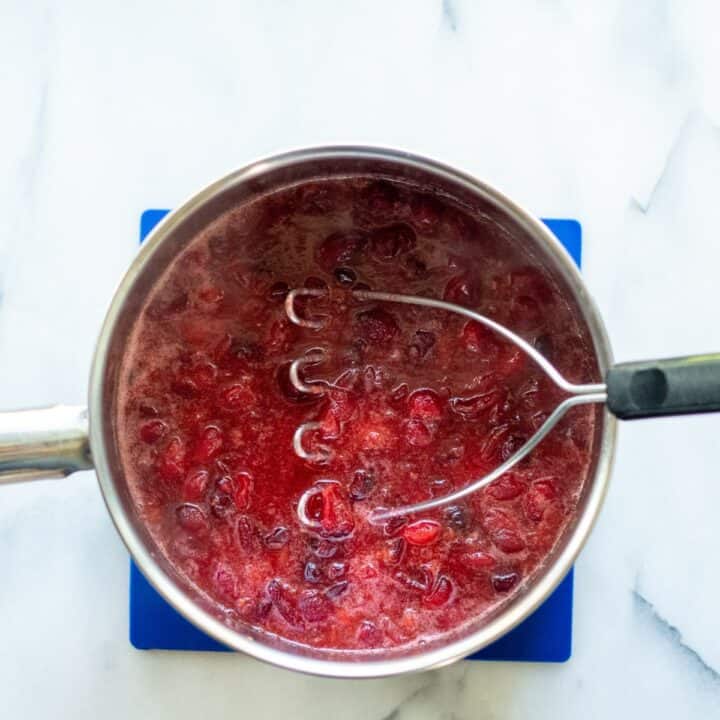 The cooked cranberries being mashed with a masher in a small pot.