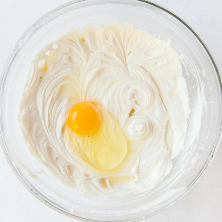 An egg being added to the mixed cream cheese and sweetener in a glass bowl.