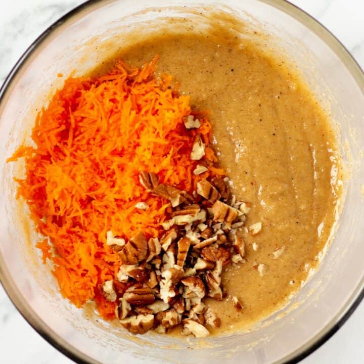 Shredded carrots and chopped pecans being added to the cake batter in a glass bowl.