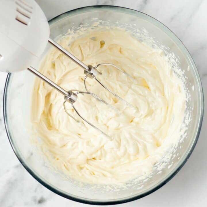 Glass bowl with cream cheese, powdered sweetener and vanilla extract being mixed with a hand mixer to make the cream cheese frosting.