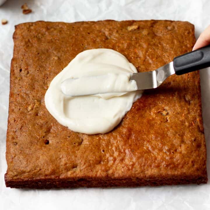 The cream cheese frosting being spread on the square carrot cake with an offset spatula.