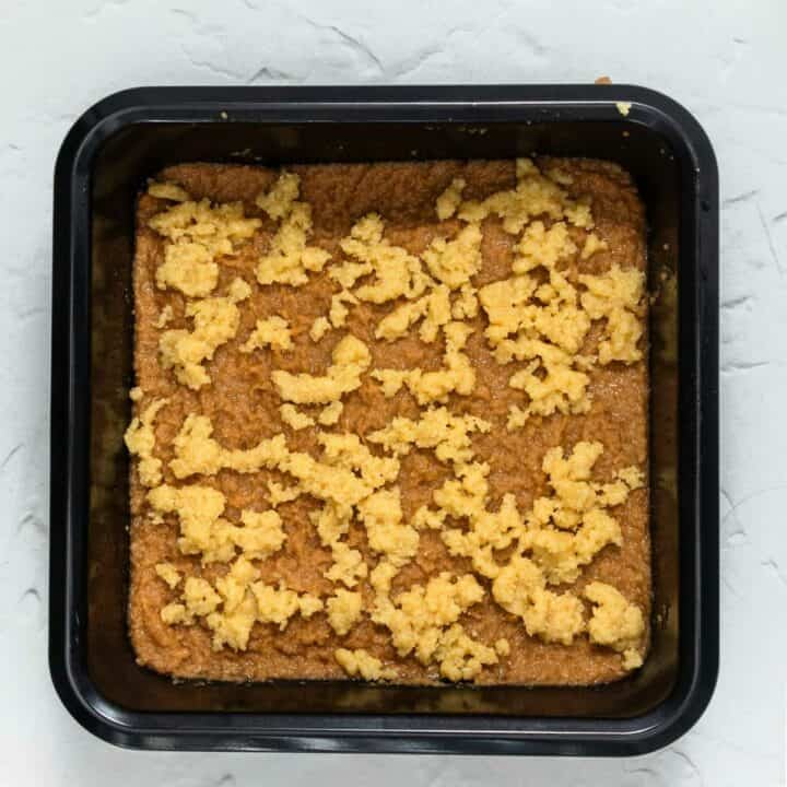 A square cake pan with keto coffee cake batter with pieces of crumb topping sprinkled on top. Ready to bake.