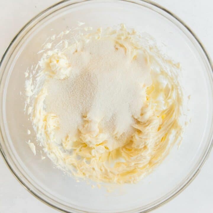 The granulated sugar being added to the mixed butter and cream cheese in a glass bowl.