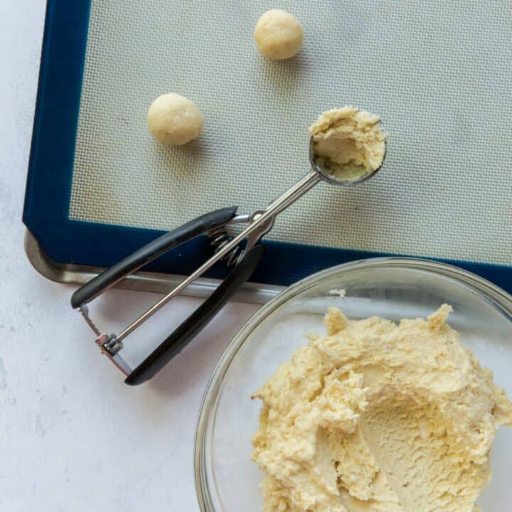 A lined baking sheet with 2 cookie dough balls and a cookie scooper with a scoop of the keto cream cheese cookie dough laying on it. A glass bowl filled with the cookie dough is next to it.