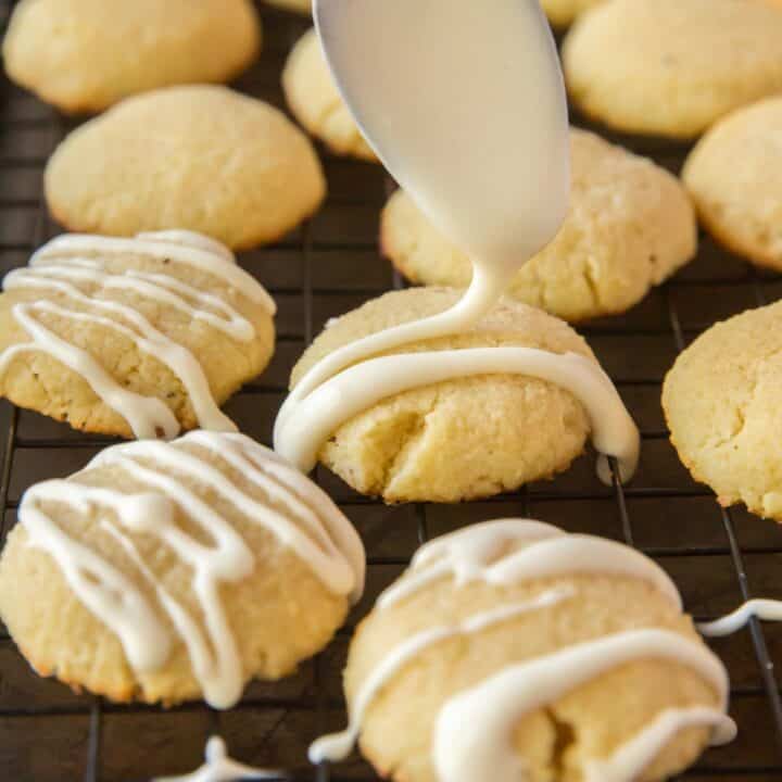 Fully baked keto cream cheese cookies on a wire cooling rack. Icing on a spoon is being drizzled on some of the cookies.