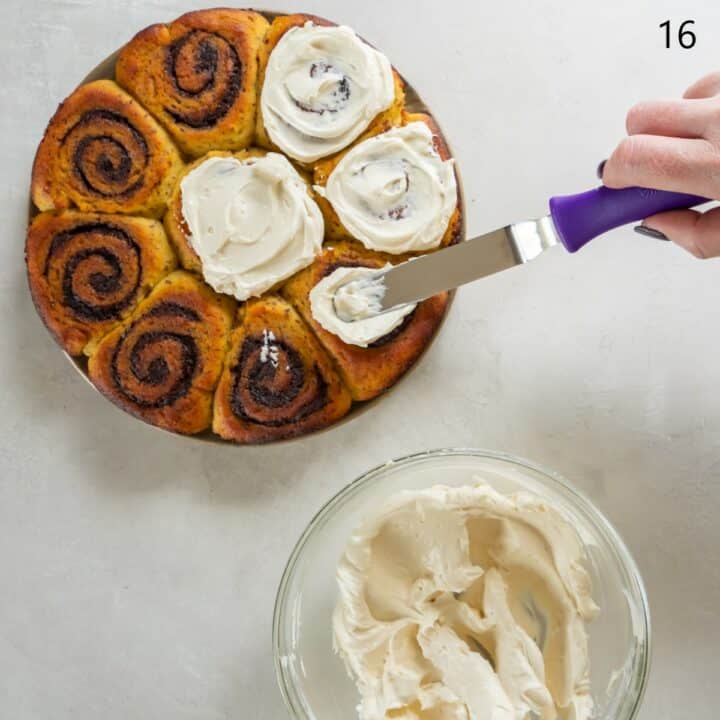 The fully baked and cooled keto cinnamon rolls, still in the cake pan, being iced with an offset spatula. A glass bowl of cream cheese frosting sits next to them.