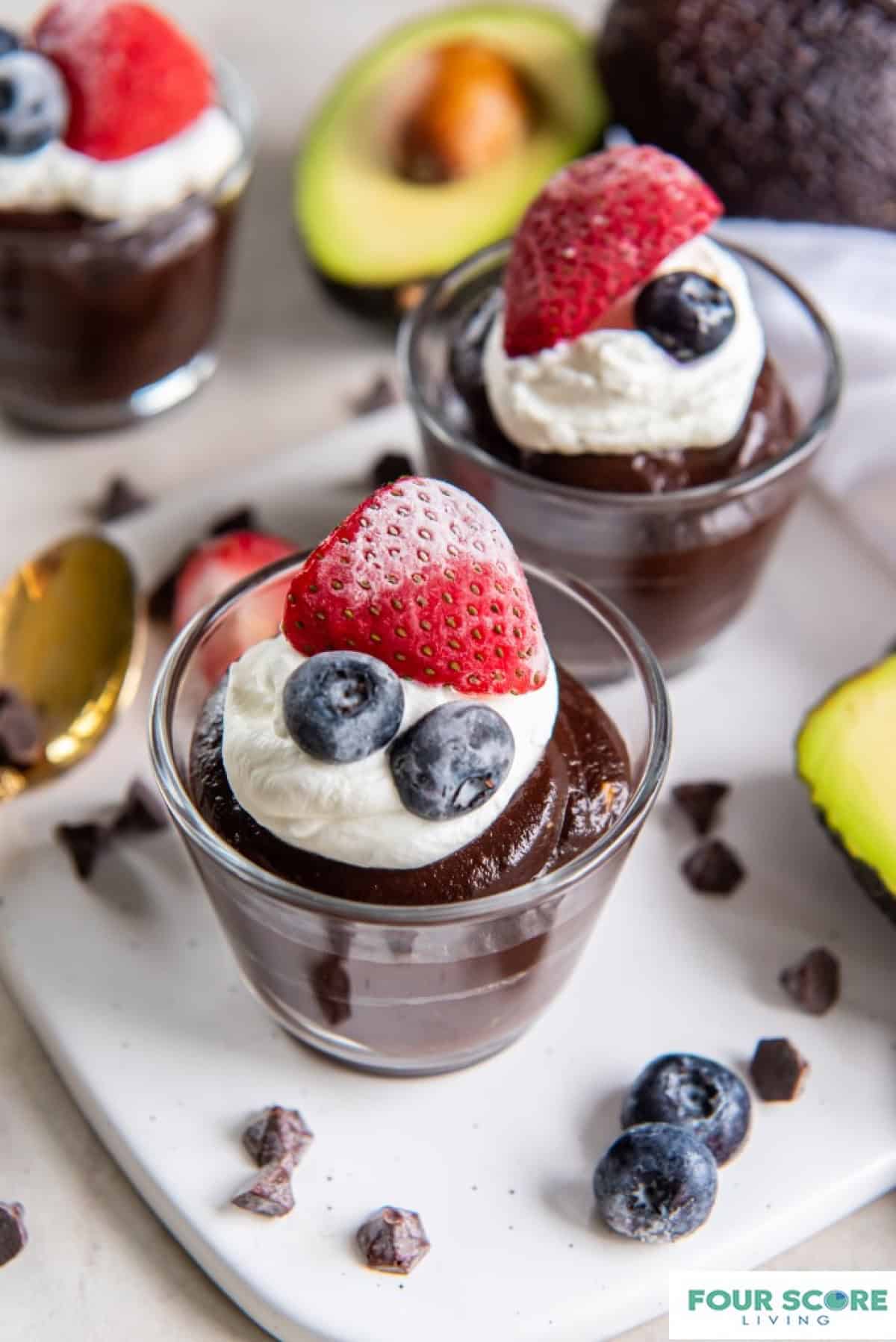 2 glass dessert cups filled with chocolate avocado pudding. The pudding is topped with a dollop of whipped cream, a half of a strawberry and two blueberries.