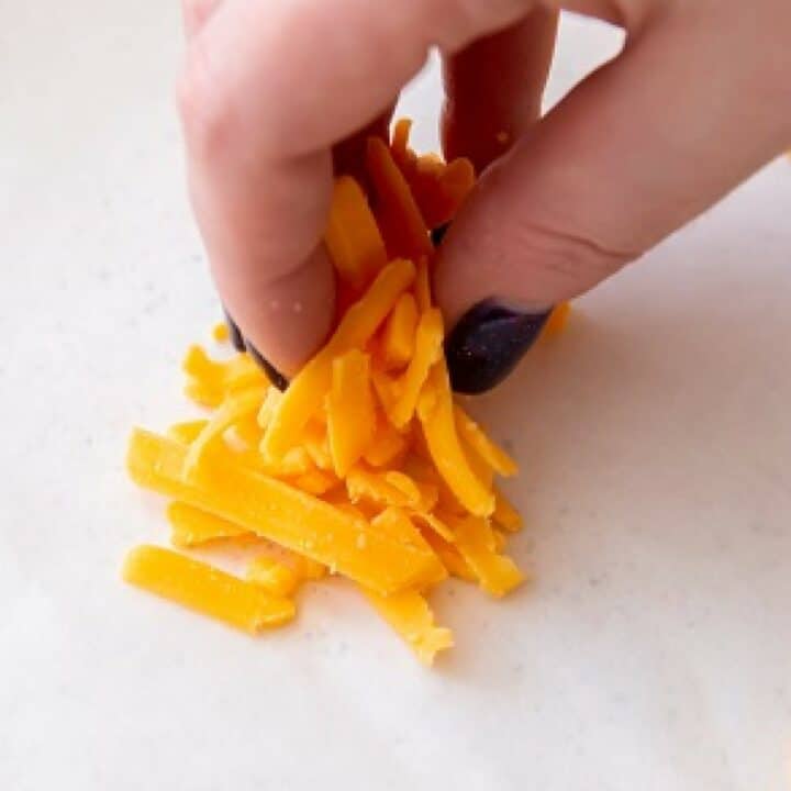 A hand making a pile of shredded cheddar cheese on a parchment lined baking sheet.