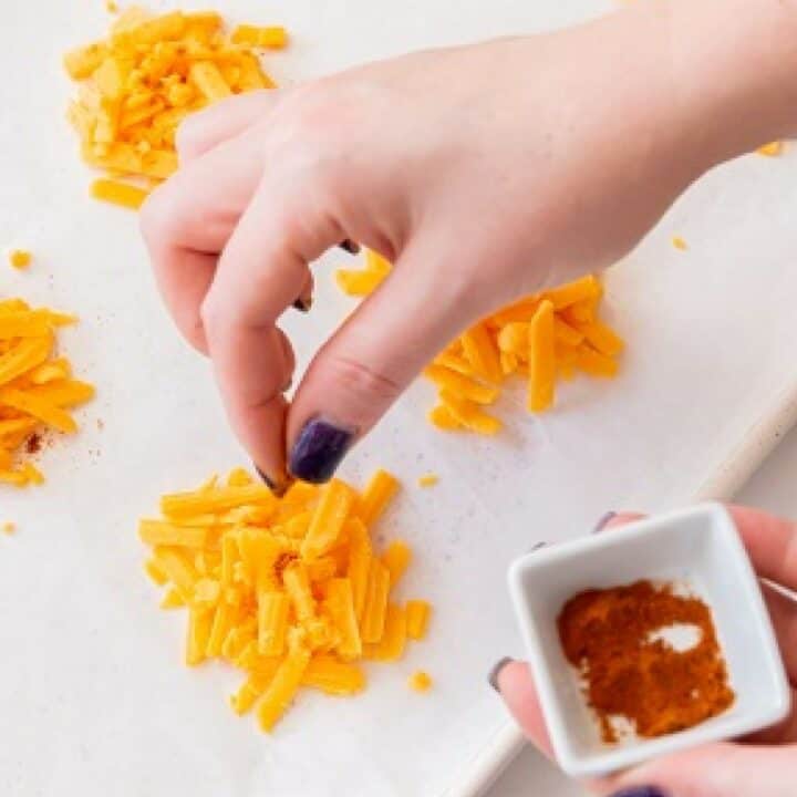 A hand sprinkling cayenne pepper on small piles of shredded cheddar cheese.