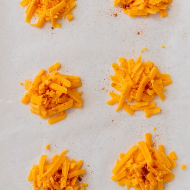 Six piles of shredded cheddar cheese with cayenne seasoning on a parchment lined baking sheet.