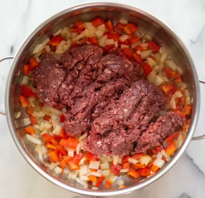 Ground beef being added to the cooked onions and bell peppers in a large pot.