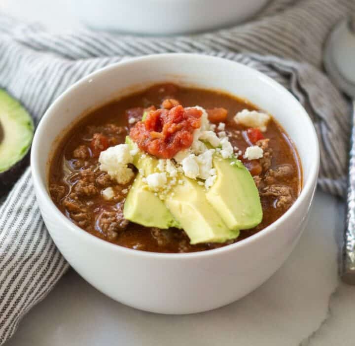 A bowl of keto taco soup with ground beef, topped with slices of avocado, salsa and crumbled cojita cheese.